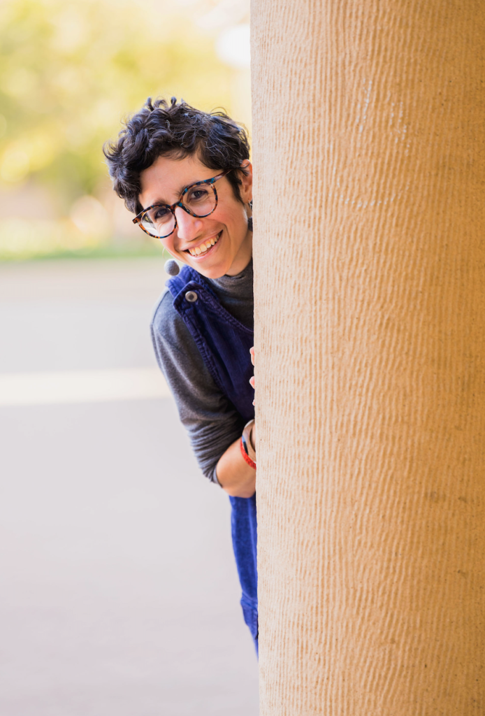 a portrait of Natalie Jabbar wearing a blue jumper and peaking behind a sandstone pillar. Photo by Harrison Truong